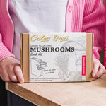 Person holding a 'Grow Your Own Mushrooms' book kit on a table with books in the background.
