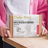 Person holding a 'Grow Your Own Mushrooms' book kit on a table with books in the background.