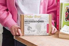 Person holding a 'Grow Your Own Mushrooms' book kit on a table with books in the background.