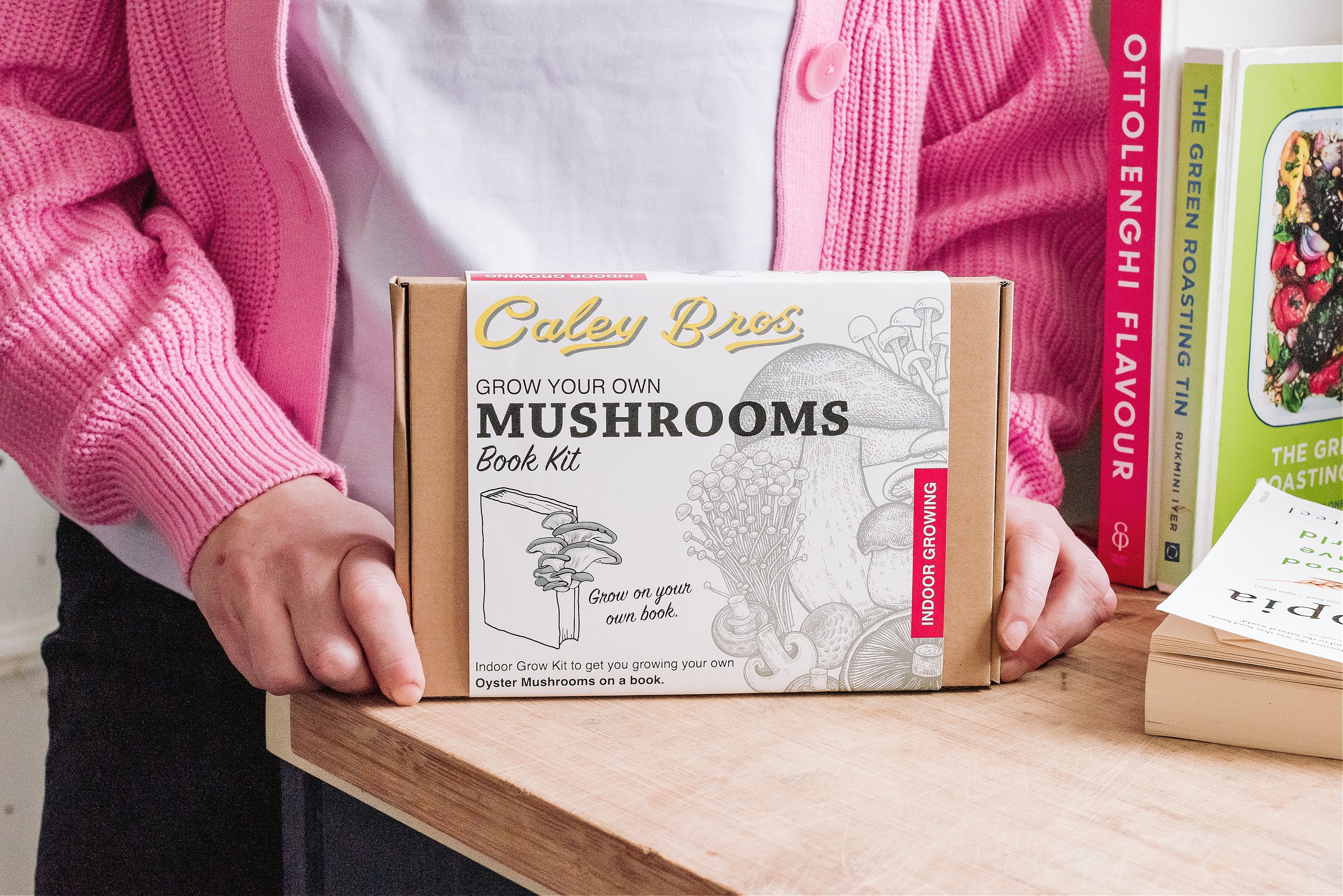 Person holding a 'Grow Your Own Mushrooms' book kit on a table with books in the background.