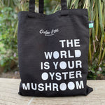 Black tote bag with white text on a wooden surface with plants in the background