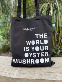 Black tote bag with white text on a wooden surface with plants in the background