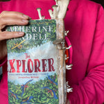 Person holding a book titled 'The Explorer' by Katherine Lundell, with a pink sweater. Mushtooms growing on book