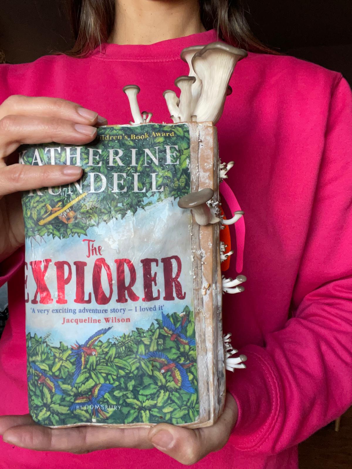 Person holding a book titled 'The Explorer' by Katherine Lundell, with a pink sweater. Mushtooms growing on book