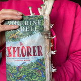 Person holding a book titled 'The Explorer' by Katherine Lundell, with a pink sweater. Mushtooms growing on book